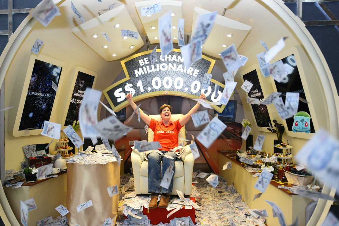 A picture of a man celebrating in front of a sign called ‘Be a Changi Millionaire’ with currency notes raining down